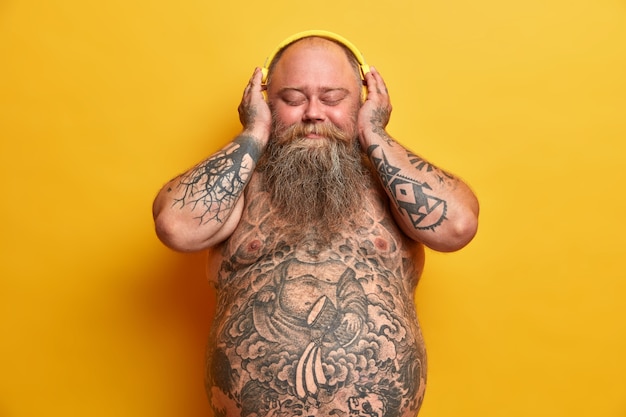 Hombre regordete complacido escucha música en auriculares con placer, cierra los ojos, se para desnudo, tiene el cuerpo tatuado, grasa que sobresale de la barriga, barba espesa, disfruta de un buen sonido, aislado en una pared amarilla