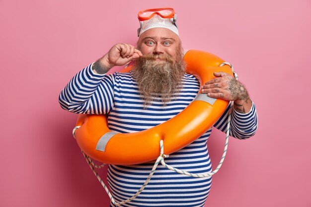 El hombre regordete positivo riza el bigote con gafas de natación y camisa de marinero a rayas, posa con equipo de seguridad en la playa, disfruta de las vacaciones de verano. Concepto de descanso y temporada