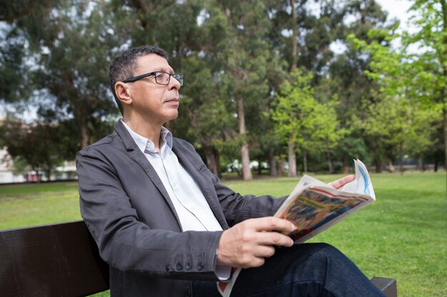 Hombre relajado que lee el periódico y que se sienta en banco en parque