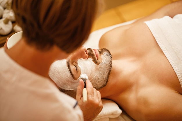 Hombre relajado recibiendo mascarilla facial durante el tratamiento cosmético en el salón de spa