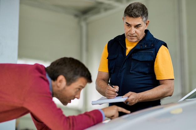 Foto gratuita hombre de reparación de automóviles escribiendo notas mientras su cliente señala un lugar problemático en el capó del vehículo en un taller