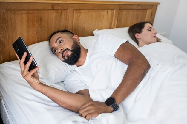 Hombre revisando su teléfono en la cama junto a su esposa