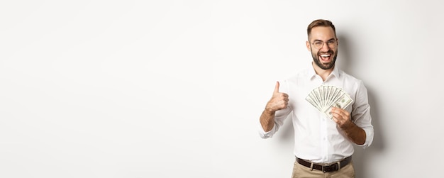 Foto gratuita hombre rico emocionado sosteniendo dinero mostrando el pulgar hacia arriba en señal de aprobación de pie sobre fondo blanco.