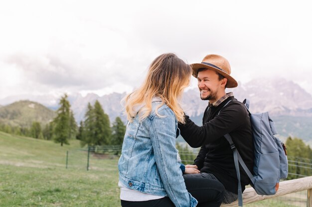 Hombre riendo con mochila azul mirando a su novia rubia sentada en el campo en la montaña