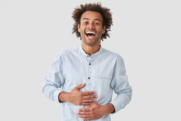 Hombre rizado de piel oscura con gran espíritu, se ríe alegremente, oye algo gracioso, vestido con una camisa elegante, posa contra la pared blanca. Un joven afroamericano satisfecho se siente muy feliz