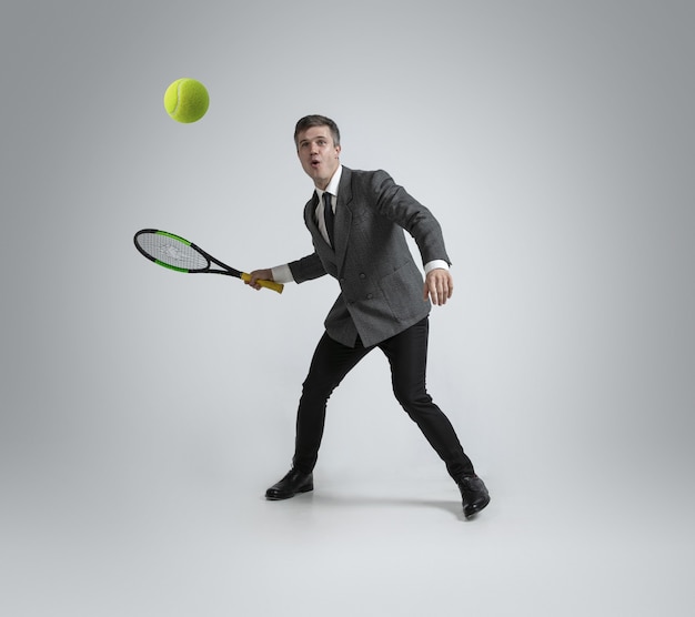 Hombre en ropa de oficina juega tenis aislado sobre fondo gris de estudio.