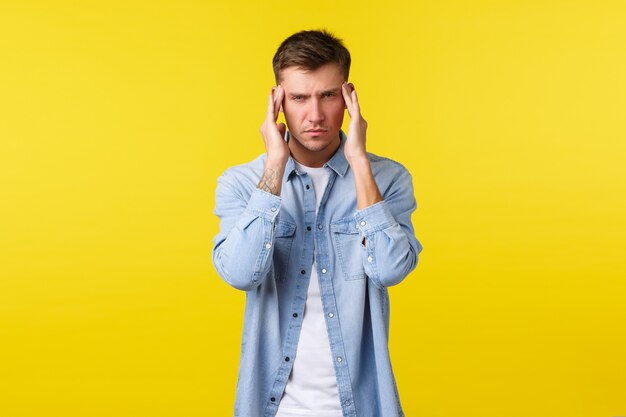 Hombre rubio guapo angustiado sintiendo estrés y fatiga. Guy sufre dolor de cabeza, toca heach y hace muecas de dolor. Hombre con migraña frotando las sienes, tratando de enfocar, fondo amarillo.