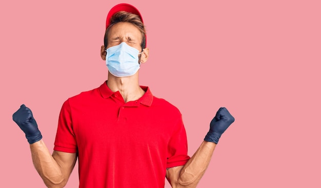 Hombre rubio guapo con barba con uniforme de entrega y máscara médica muy feliz y emocionado haciendo gesto de ganador con los brazos levantados sonriendo y gritando por el concepto de celebración de éxito