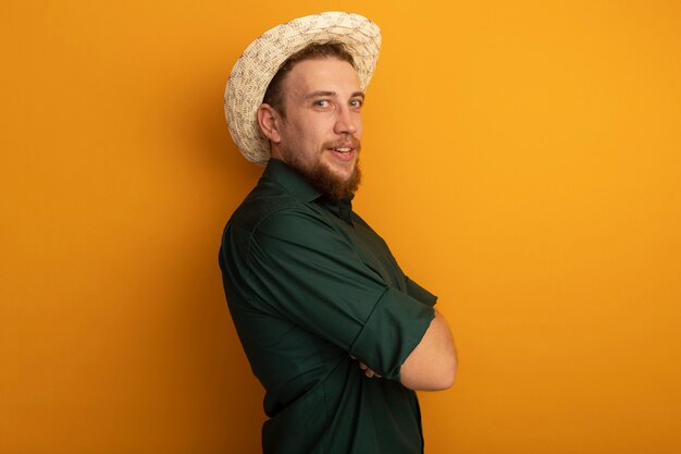 Hombre rubio guapo complacido con sombrero de playa se encuentra de lado con los brazos cruzados aislados en la pared naranja