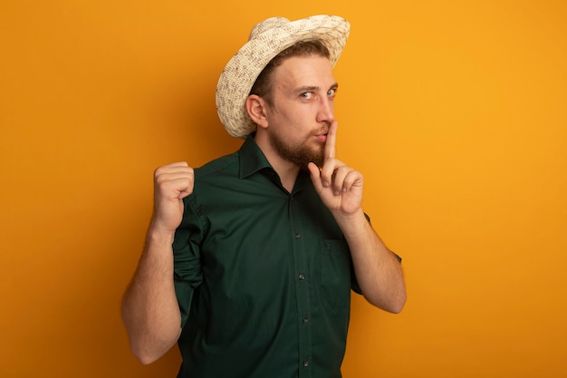 Hombre rubio guapo complacido con sombrero de playa haciendo gesto de silencio y mantiene el puño aislado en la pared naranja