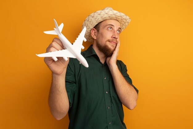 Foto gratuita hombre rubio guapo complacido con sombrero de playa pone la mano en la cara y sostiene el avión modelo aislado en la pared naranja