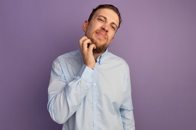 Hombre rubio guapo disgustado rascarse la barba aislada en la pared púrpura