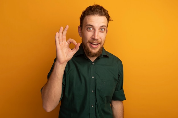 Hombre rubio guapo emocionado gestos ok signo de mano aislado en la pared naranja