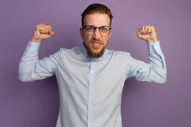 Hombre rubio guapo molesto con gafas ópticas mantiene los puños aislados en la pared púrpura