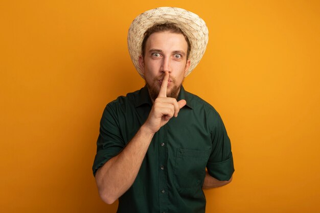 Foto gratuita hombre rubio guapo sorprendido con sombrero de playa haciendo gesto de silencio en naranja