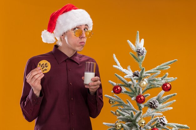 Hombre rubio joven confundido con sombrero de santa y gafas de pie cerca del árbol de Navidad decorado con vaso de leche y galletas mirando a cámara aislada sobre fondo naranja