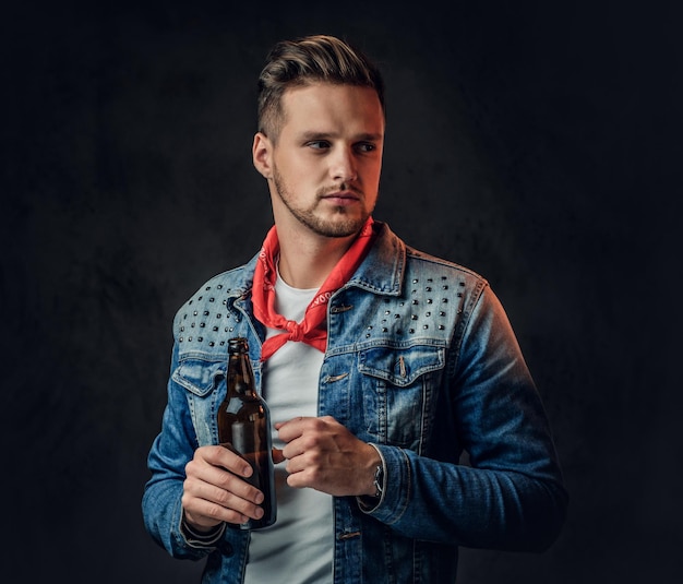 Foto gratuita un hombre rubio positivo vestido con una chaqueta vaquera sostiene una botella con cerveza artesanal.