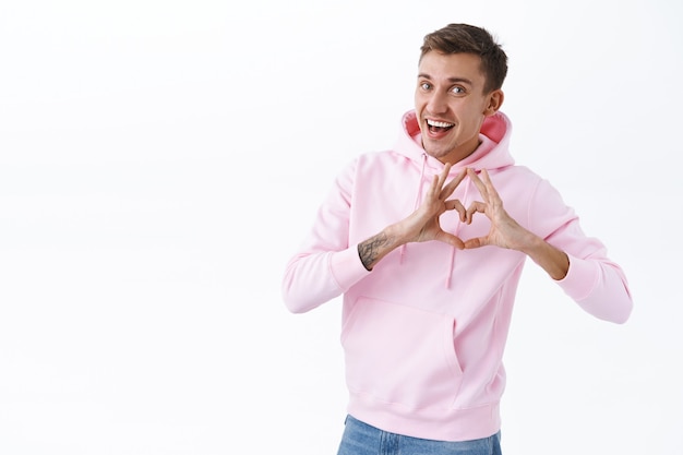 El hombre rubio romántico alegre expresa su simpatía, felicita al amante con el aniversario, muestra el signo del corazón mientras confiesa el amor y los sentimientos tiernos, sonríe optimista, de pie en la pared blanca