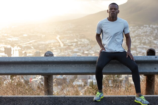 Hombre sano con piel negra, descansa en la señal de tráfico, viste ropa casual, mira con confianza a la cámara, modela contra la vista panorámica con montañas y ciudad, lleva un estilo de vida saludable. Entrenamiento físico