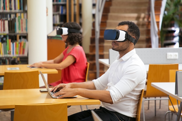 Hombre seguro escribiendo en la computadora portátil mientras está sentado en un auricular VR