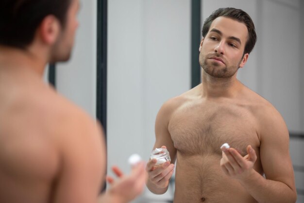 Hombre semidesnudo de pie cerca del espejo y aplicando espuma de afeitar en la cara