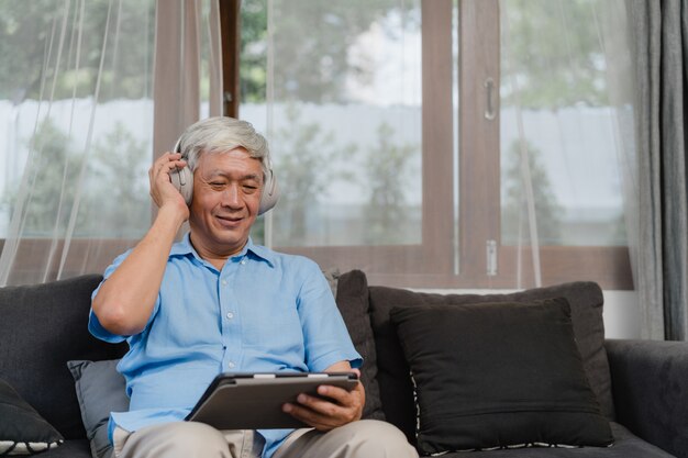 Hombre senior asiático relajarse en casa. Auriculares felices masculinos más viejos asiáticos del desgaste usando el podcast que escucha de la tableta mientras que miente en el sofá en concepto de la sala de estar en casa.