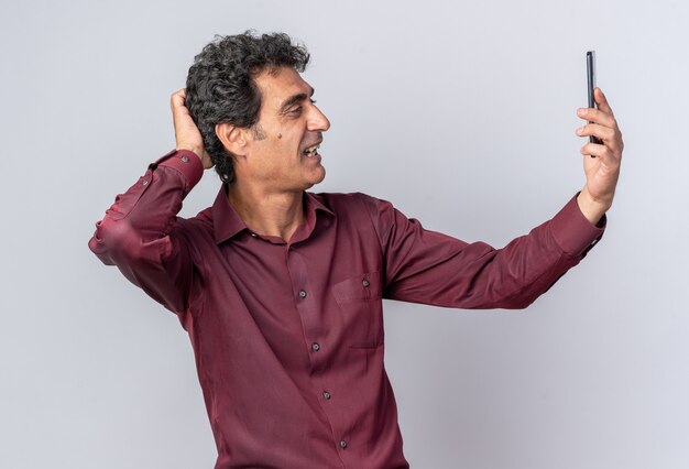 Hombre senior en camisa púrpura haciendo selfie con smartphone feliz y positivo sonriendo alegremente