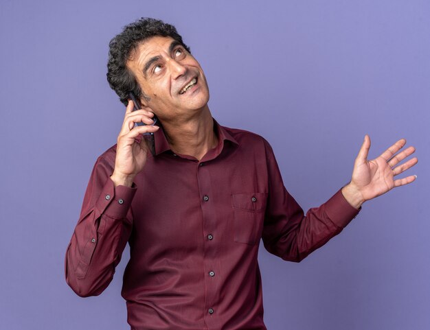 Hombre senior en camisa púrpura mirando hacia arriba sonriendo alegremente mientras habla por teléfono móvil de pie sobre fondo azul.