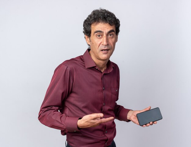 Hombre senior en camisa púrpura mirando confundido sosteniendo smartphone parado sobre blanco