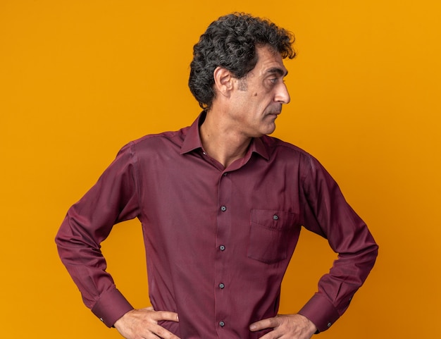 Foto gratuita hombre senior en camisa púrpura mirando a un lado con cara seria de pie sobre fondo naranja