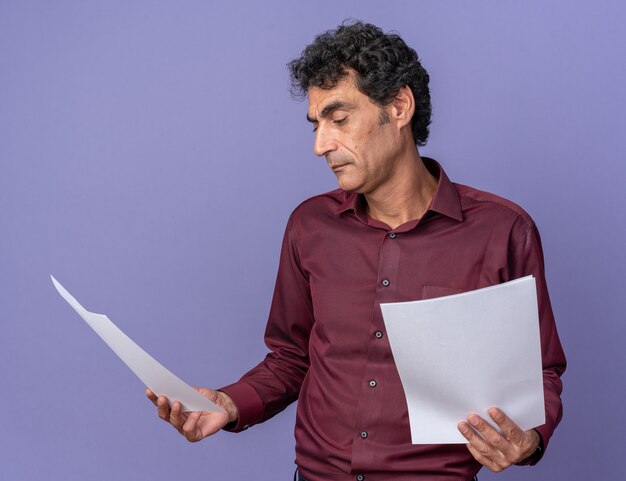 Hombre senior en camisa púrpura sosteniendo páginas en blanco mirando confundido teniendo dudas sobre azul