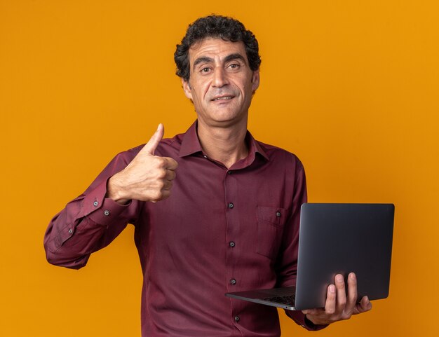 Hombre senior en camisa púrpura sosteniendo el portátil mirando a la cámara sonriendo confiado mostrando Thumbs up parado sobre naranja