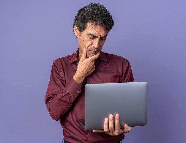 Hombre senior en camisa púrpura sosteniendo portátil mirando la pantalla desconcertado
