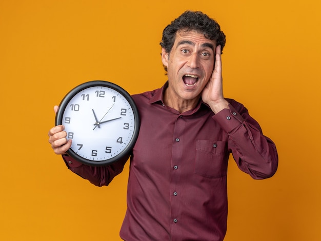 Hombre senior en camisa púrpura sosteniendo el reloj de pared mirando a la cámara asombrado y sorprendido de pie sobre fondo naranja