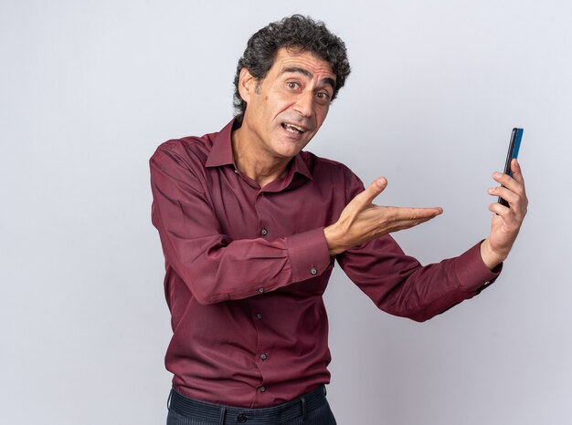 Hombre senior en camisa púrpura sosteniendo smartphone presentando con el brazo de la mano feliz y positivo