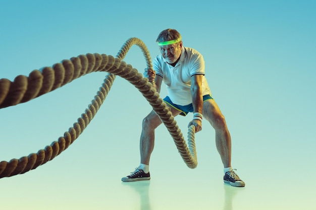 Hombre senior entrenando con cuerdas sobre fondo degradado en luz de neón. Modelo masculino caucásico en gran forma se mantiene activo, deportivo.