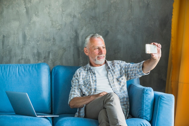 Hombre senior feliz sentado en el sofá azul tomando selfie en teléfono móvil