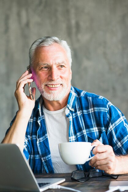 Hombre senior feliz sosteniendo la taza de café hablando por teléfono inteligente