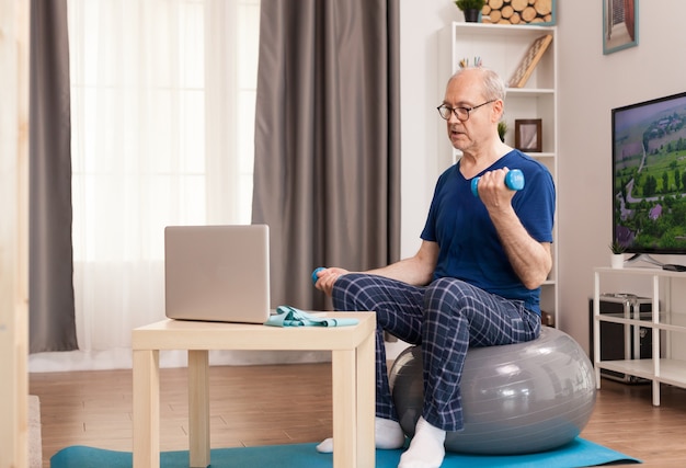 Hombre senior de formación con entrenador en línea sentado sobre una pelota suiza en el medio de la habitación