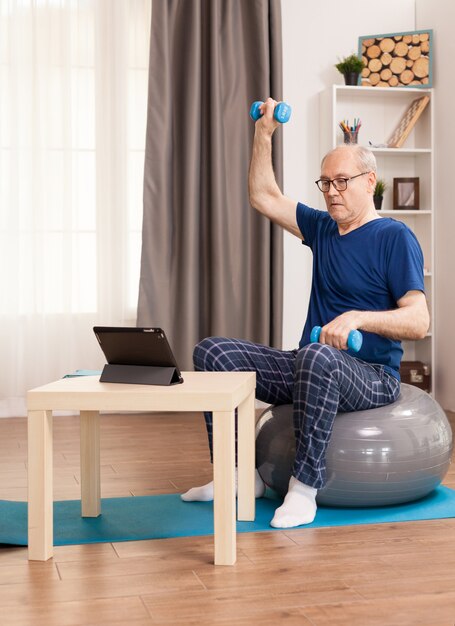 Hombre senior haciendo ejercicios de recuperación muscular sentado sobre una pelota suiza frente a la tableta