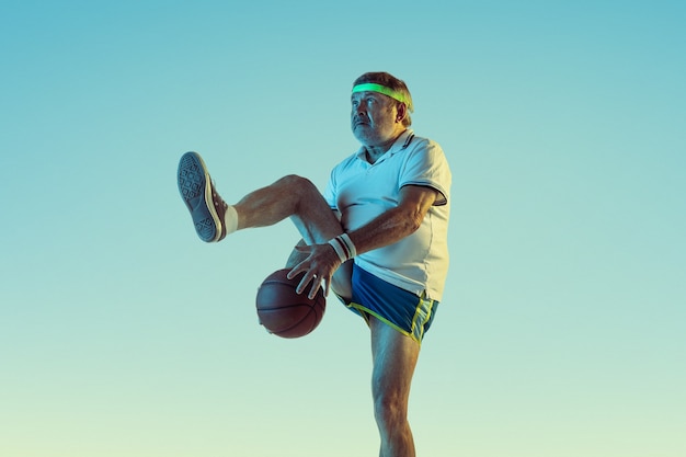 Hombre senior jugando baloncesto en pared degradada con luz de neón. Modelo masculino caucásico en gran forma se mantiene activo, deportivo. Concepto de deporte, actividad, movimiento, bienestar, estilo de vida saludable.