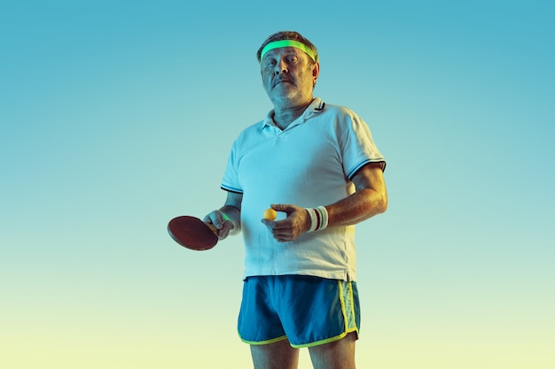 Hombre senior jugando tenis de mesa en pared degradada en luz de neón. Modelo masculino caucásico en gran forma se mantiene activo, deportivo. Concepto de deporte, actividad, movimiento, bienestar, estilo de vida saludable.