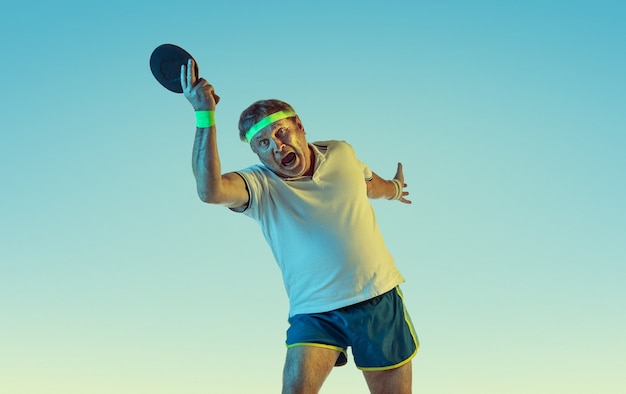 Hombre senior jugando tenis de mesa en pared degradada en luz de neón. Modelo masculino caucásico en gran forma se mantiene activo, deportivo. Concepto de deporte, actividad, movimiento, bienestar, estilo de vida saludable.