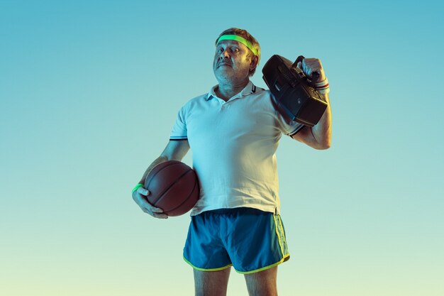 Hombre senior posando impresionante en ropa deportiva con grabadora retro en pared degradada, neón. Modelo masculino caucásico en gran forma, deportivo. Concepto de deporte, actividad, movimiento, estilo de vida saludable.