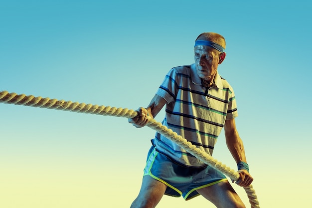 Foto gratuita hombre senior con ropa deportiva entrenando con cuerdas sobre fondo degradado, luz de neón.