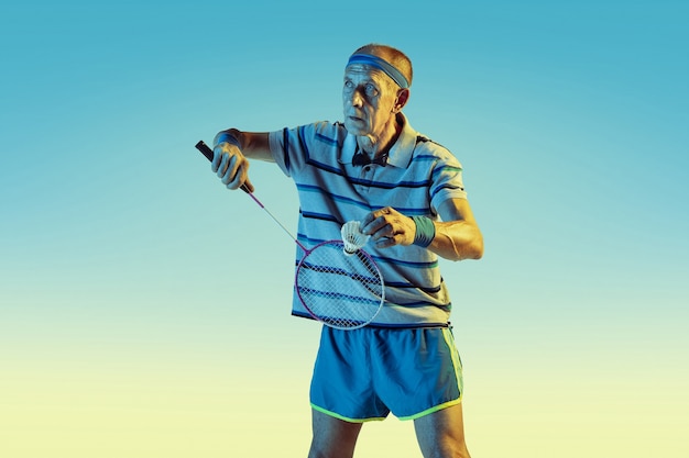 Hombre senior con ropa deportiva jugando bádminton sobre fondo degradado, luz de neón. Modelo masculino caucásico en gran forma se mantiene activo. Concepto de deporte, actividad, movimiento, bienestar, confianza.