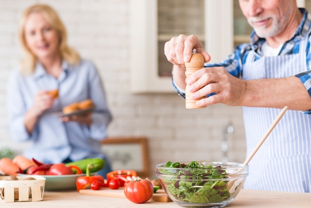 Hombre senior sazonando la ensalada de verduras verdes y su esposa sosteniendo los muffins en la mano en el fondo