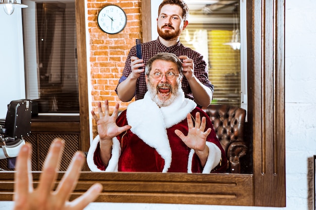 Hombre senior en traje de Santa Claus afeitando a su maestro personal en la peluquería antes de Navidad