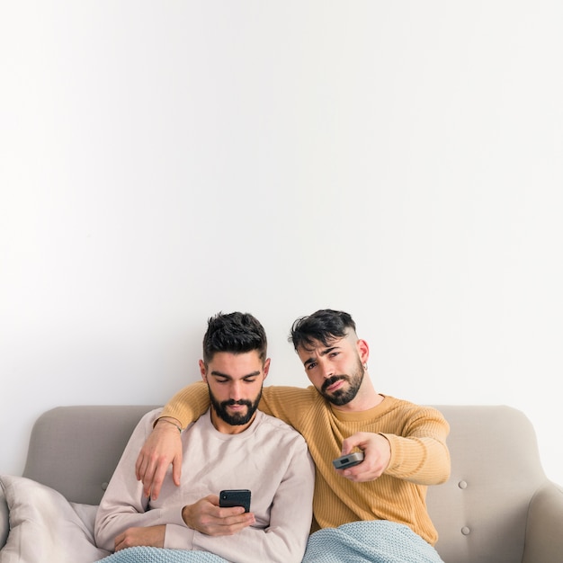 Hombre sentado al lado de su novio usando el teléfono móvil cambiando el canal de televisión con control remoto