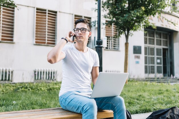 Hombre sentado en la banca con la computadora portátil hablando por teléfono móvil